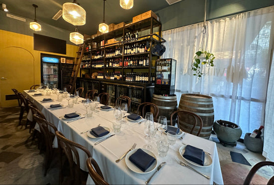 Dining area with a long table set for dinner, wine bottles on shelves, and large windows.